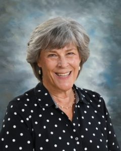 Smiling woman with gray hair, wearing a black polka dot blouse, standing against a cloudy background.