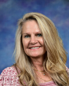 Smiling woman with long blonde hair, wearing a patterned top, against a blue background.
