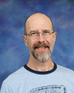 A smiling man with glasses and a beard wears a light blue shirt, set against a soft blue background.