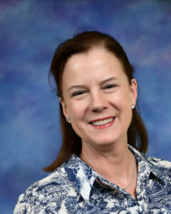 A woman with brown hair smiles warmly, wearing a patterned blouse, against a soft blue background.