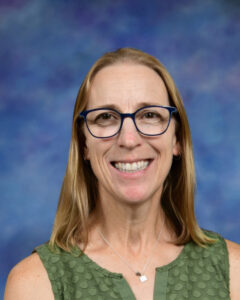 Smiling woman with blonde hair and glasses, wearing a green top, against a blue abstract background.