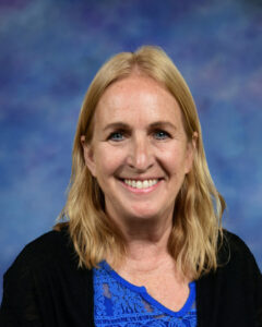 Smiling woman with long blonde hair, wearing a blue top and black cardigan, against a colorful background.