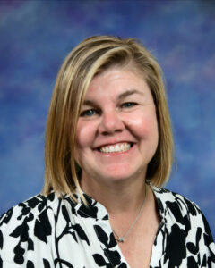 Smiling woman with blonde hair, wearing a black and white floral shirt, against a blue background.