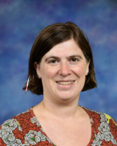 A smiling woman with short brown hair, wearing a patterned top, set against a blue background.