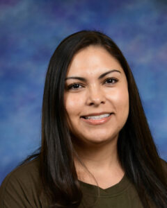A woman with long dark hair smiles in front of a colorful blue background, wearing a brown shirt.