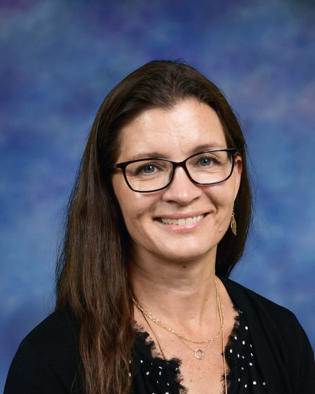 A woman with long brown hair and glasses smiles, wearing a black top against a blue and purple background.