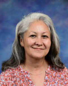 A smiling woman with long, gray hair wearing a floral blouse, set against a blue background.