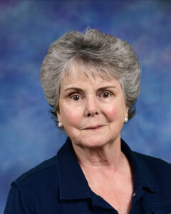 A senior woman with short gray hair, wearing a navy blue shirt and pearl earrings, against a blue background.
