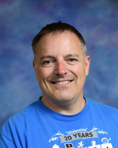 Smiling man with short hair wearing a blue t-shirt, against a colorful blue background.