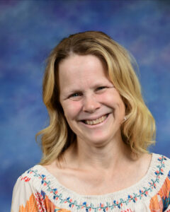 Smiling woman with long, wavy blonde hair wearing a colorful, floral-patterned top against a blue background.