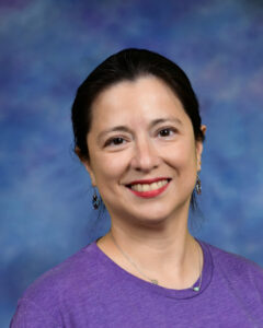 A woman with dark hair smiles in a purple shirt, against a blurred blue background. She wears earrings and a necklace.