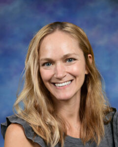 Smiling woman with long, wavy blonde hair, wearing a gray shirt, against a soft blue background.