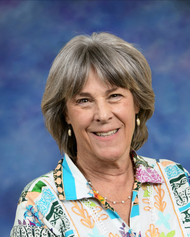 Smiling woman with short gray hair, wearing a colorful patterned shirt, against a blue background.