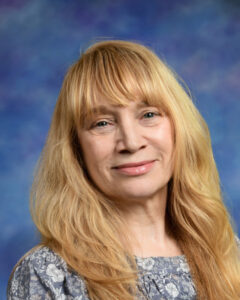 A woman with long, wavy blonde hair smiles, wearing a floral-patterned top against a blue background.