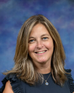 A woman with long brown hair smiles, wearing a black top with ruffled sleeves, against a blue background.