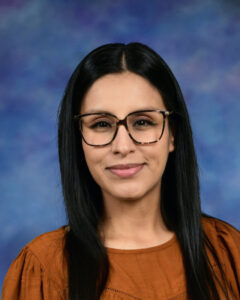 A woman with long black hair and glasses smiles, wearing an orange top against a blue background.
