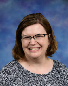 Smiling woman with shoulder-length brown hair, wearing glasses and a floral top, against a blue background.