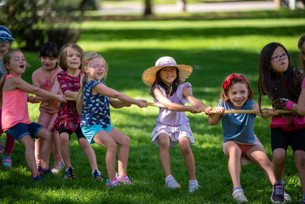 Seven elementary school-aged girls laughing while pulling on a tug-of-war rope in the grass. 