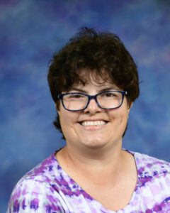 Persona sonriente con pelo corto oscuro y gafas, vistiendo una camisa con motivos morados, sobre un fondo azul.