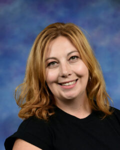 Smiling woman with wavy blonde hair wearing a black shirt, set against a blue background.