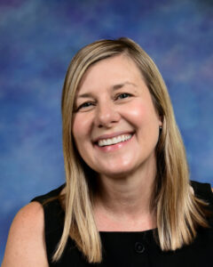 A smiling woman with long, straight blonde hair, wearing a black top, against a blue and purple background.