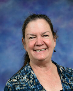 Smiling woman with long hair wearing a blue patterned shirt, set against a soft purple background.