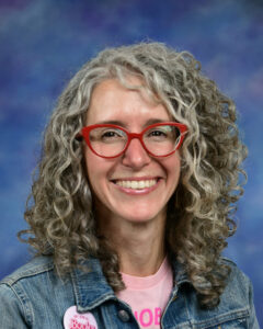 Smiling woman with curly gray hair and red glasses, wearing a denim jacket and a pink shirt with a badge.