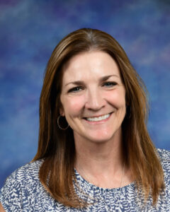Smiling woman with long brown hair, wearing a patterned shirt, against a blue and purple background.